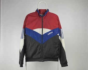 Puede incluir: Chaqueta deportiva Adidas vintage en rojo, negro, azul y blanco. La chaqueta tiene cremallera completa, cuello alto y el logo de Adidas en el pecho. Las mangas tienen rayas blancas.