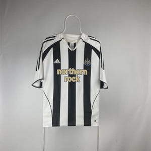 Pu&ograve; includere: Maglia da calcio a righe bianche e nere con la scritta "northern rock" in oro. La maglia ha uno scollo a V, il logo Adidas e lo stemma del Newcastle United. &Egrave; appesa.