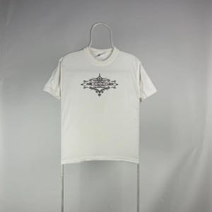 May include: A white t-shirt with a crew neck and short sleeves. The shirt features a black decorative design with the word "INDEPENDENT" in the center. The shirt is hanging on a metal hanger.
