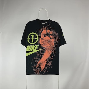 May include: Black Nike t-shirt with a large orange footprint graphic and the Nike logo in neon green. The shirt also features a circular design with the number 1 and a blueprint-style background.