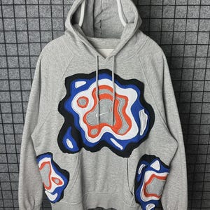 Puede incluir: Sudadera con capucha gris con cordón blanco y un gran diseño abstracto en azul, naranja y negro en la parte delantera. Diseños abstractos adicionales están en el bolsillo y las mangas. La sudadera tiene un bolsillo de canguro y una capucha forrada en blanco.