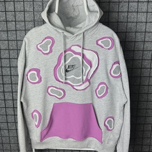 Puede incluir: Sudadera con capucha gris con cordón blanco y bolsillo de canguro morado vibrante. La sudadera presenta un diseño único con formas superpuestas moradas y blancas, y el logo de Nike.