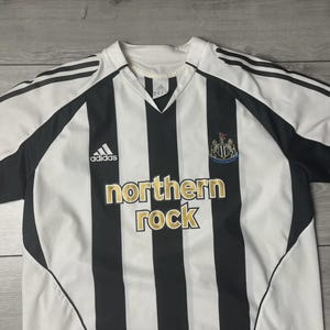 Pu&ograve; includere: Maglia da calcio a righe verticali bianche e nere. Presenta il logo Adidas, lo stemma del club e la scritta "northern rock" in lettere dorate. Scollo a V e maniche nere.