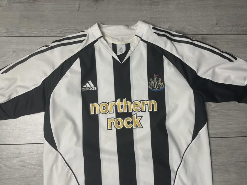 Pu&ograve; includere: Maglia da calcio a righe verticali bianche e nere. Presenta il logo Adidas, lo stemma del club e la scritta "northern rock" in lettere dorate. Scollo a V e maniche nere.