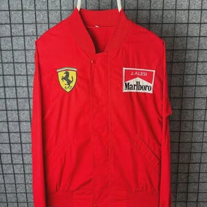 May include: Bright red bomber jacket with the Ferrari logo and a Marlboro patch. The jacket has a button closure and two front pockets. The jacket is hanging on a white hanger.