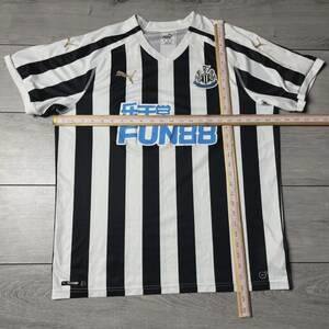 Puede incluir: Una camiseta de f&uacute;tbol a rayas blancas y negras con cuello en V. La camiseta presenta el texto "FUN88" en azul, un logotipo del equipo y el logotipo de Puma en dorado. La camiseta est&aacute; sobre una superficie de madera.