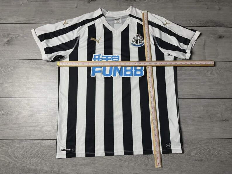 Puede incluir: Una camiseta de f&uacute;tbol a rayas blancas y negras con cuello en V. La camiseta presenta el texto "FUN88" en azul, un logotipo del equipo y el logotipo de Puma en dorado. La camiseta est&aacute; sobre una superficie de madera.