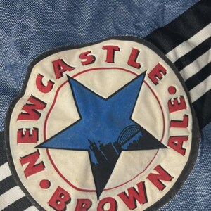 K&ouml;nnte beinhalten: Nahaufnahme eines Newcastle Brown Ale-Aufn&auml;hers. Der Aufn&auml;her zeigt einen blauen Stern mit einer Silhouette einer Br&uuml;cke und Geb&auml;uden, umgeben von einem roten Kreis mit den Worten "NEWCASTLE BROWN ALE". Der Hintergrund ist blau.