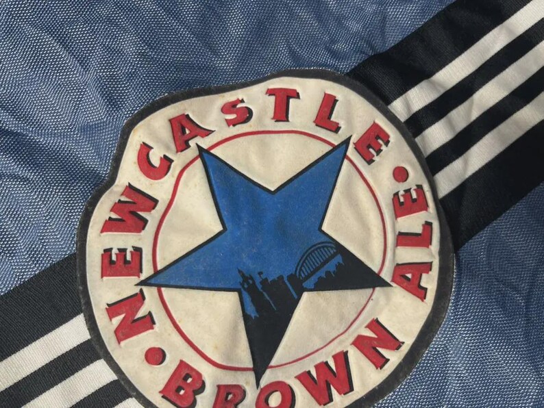 K&ouml;nnte beinhalten: Nahaufnahme eines Newcastle Brown Ale-Aufn&auml;hers. Der Aufn&auml;her zeigt einen blauen Stern mit einer Silhouette einer Br&uuml;cke und Geb&auml;uden, umgeben von einem roten Kreis mit den Worten "NEWCASTLE BROWN ALE". Der Hintergrund ist blau.