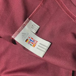 May include: Close-up of a maroon athletic garment with a stitched seam. A white tag with the Adidas logo and international flags is visible. The tag also includes the numbers "07/03" and "021778".