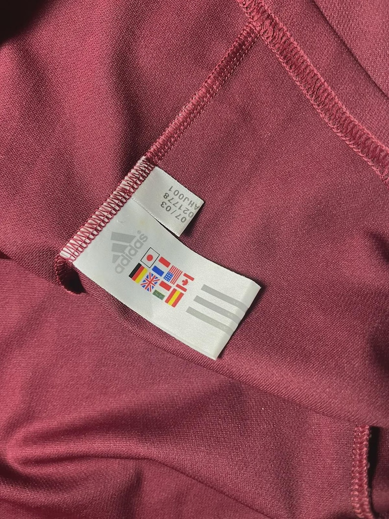 May include: Close-up of a maroon athletic garment with a stitched seam. A white tag with the Adidas logo and international flags is visible. The tag also includes the numbers "07/03" and "021778".