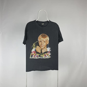 May include: Black t-shirt featuring a portrait of a young man with blonde hair, a signature, and the name "NICK" in a stylized font. The shirt is hanging on a white hanger against a plain background. The shirt appears to be vintage.