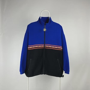 May include: A blue and black zip-up fleece jacket with a high collar. The jacket features a decorative band of orange, yellow, and red geometric patterns across the chest. The jacket is hanging on a white hanger.
