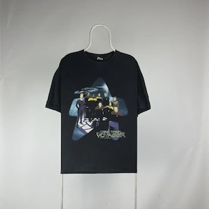 Puede incluir: Camiseta negra con un gráfico del equipo de Star Trek: Voyager dentro de un diseño en forma de estrella. El título del programa está en letras doradas. La camiseta está colgada de una percha blanca sobre un fondo liso.