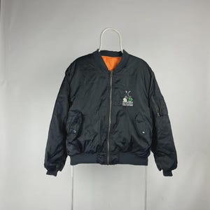Peut inclure: Veste de bombardier noire avec une doublure orange. Elle est dotée d'une fermeture éclair, de deux poches avant et d'un petit logo brodé sur la poitrine. La veste est suspendue à un cintre blanc.