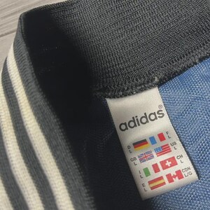K&ouml;nnte beinhalten: Nahaufnahme eines Adidas-Etiketts auf einem blau-schwarzen Kleidungsst&uuml;ck. Das Etikett zeigt das Adidas-Logo und Gr&ouml;&szlig;eninformationen sowie L&auml;nderflaggen. Das Kleidungsst&uuml;ck hat wei&szlig;e Streifen.