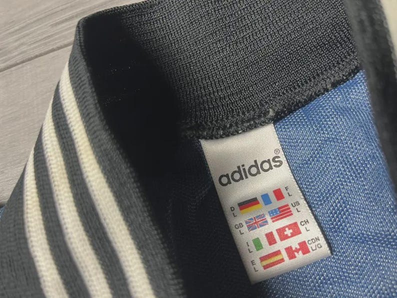 K&ouml;nnte beinhalten: Nahaufnahme eines Adidas-Etiketts auf einem blau-schwarzen Kleidungsst&uuml;ck. Das Etikett zeigt das Adidas-Logo und Gr&ouml;&szlig;eninformationen sowie L&auml;nderflaggen. Das Kleidungsst&uuml;ck hat wei&szlig;e Streifen.