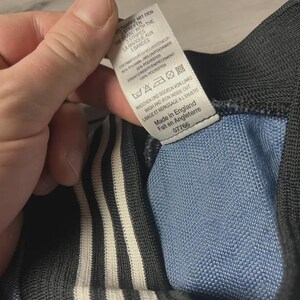 K&ouml;nnte beinhalten: Nahaufnahme eines Kleidungsst&uuml;cks mit blauem und schwarzem Design und wei&szlig;en Streifen. Ein Etikett mit englischem und franz&ouml;sischem Text ist sichtbar, das angibt, dass der Artikel in England hergestellt wurde.