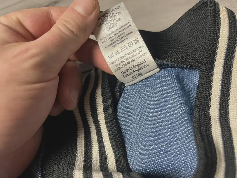 K&ouml;nnte beinhalten: Nahaufnahme eines Kleidungsst&uuml;cks mit blauem und schwarzem Design und wei&szlig;en Streifen. Ein Etikett mit englischem und franz&ouml;sischem Text ist sichtbar, das angibt, dass der Artikel in England hergestellt wurde.