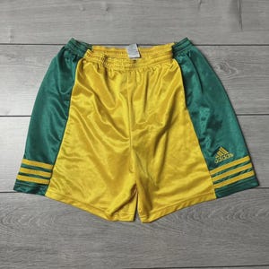 May include: Yellow and green athletic shorts with the Adidas logo. The shorts have a yellow front and green sides. The green sides have three yellow stripes near the bottom. The shorts are made of a shiny material.