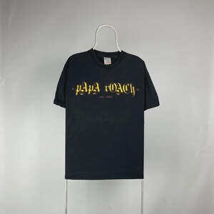 Può includere: T-shirt nera con il nome della band "Papa Roach" in lettere gialle e rosse. Il testo "est. 1993" è in basso. La maglietta è appesa a una gruccia bianca su uno sfondo neutro.