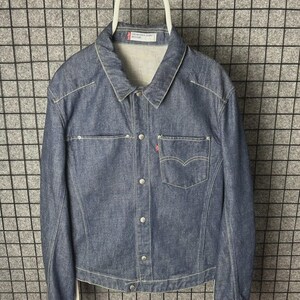 Puede incluir: Chaqueta vaquera azul con cuello clásico, cierre de botones y dos bolsillos en el pecho. La chaqueta presenta costuras blancas y una pequeña etiqueta roja en el bolsillo del pecho. Cuelga de una percha blanca.