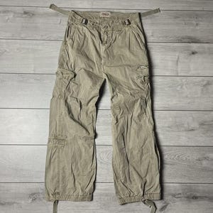 Puede incluir: Pantalones cargo de color caqui con múltiples bolsillos y correas ajustables. Los pantalones tienen un corte holgado y presentan la marca "ONLY" en la cintura. Los pantalones están colocados sobre una superficie de madera.