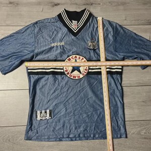 K&ouml;nnte beinhalten: Ein blaues Kurzarm-Trikot mit V-Ausschnitt, mit dem Adidas-Logo und einem Newcastle Brown Ale-Aufn&auml;her. Das Trikot hat schwarz-wei&szlig;e Streifen und ein Newcastle United-Wappen. Das Trikot liegt flach auf einer Holzoberfl&auml;che.
