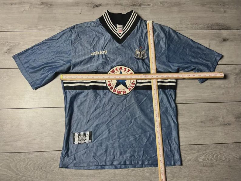 K&ouml;nnte beinhalten: Ein blaues Kurzarm-Trikot mit V-Ausschnitt, mit dem Adidas-Logo und einem Newcastle Brown Ale-Aufn&auml;her. Das Trikot hat schwarz-wei&szlig;e Streifen und ein Newcastle United-Wappen. Das Trikot liegt flach auf einer Holzoberfl&auml;che.