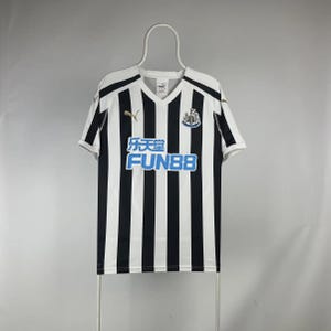 Puede incluir: Camiseta de f&uacute;tbol a rayas blancas y negras con cuello en V. La camiseta presenta el texto "乐天堂 FUN88" en azul, un logotipo de Puma y un escudo del equipo. La camiseta est&aacute; colgada de una percha blanca.