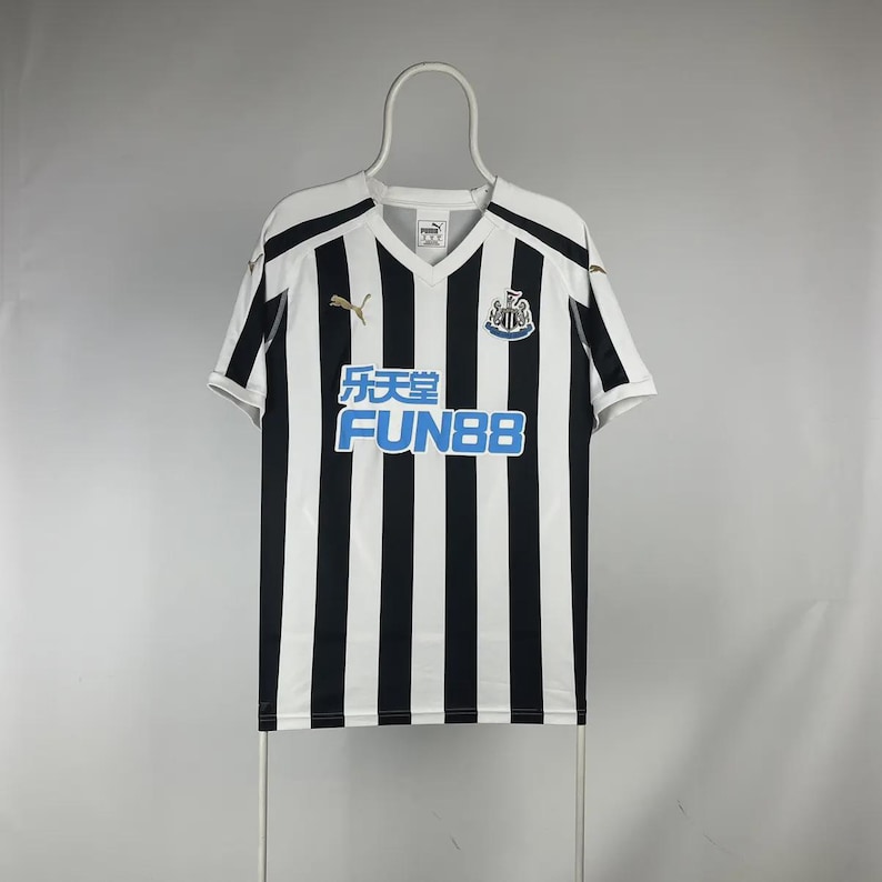 Puede incluir: Camiseta de f&uacute;tbol a rayas blancas y negras con cuello en V. La camiseta presenta el texto "乐天堂 FUN88" en azul, un logotipo de Puma y un escudo del equipo. La camiseta est&aacute; colgada de una percha blanca.