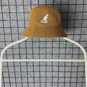 Puede incluir: Un sombrero de pescador marr&oacute;n con un logotipo de canguro blanco y la palabra "KANGOL" bordada en la parte delantera. El sombrero est&aacute; hecho de una tela texturizada y tiene un ala ligeramente inclinada. El sombrero se muestra en una percha blanca.