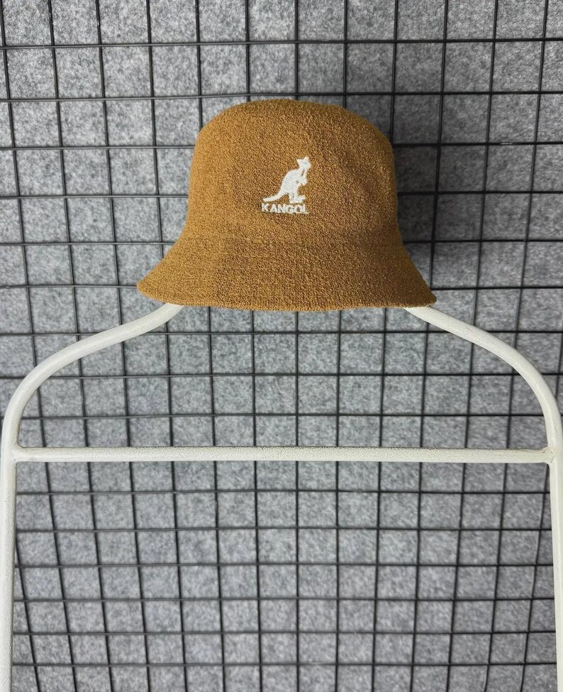 Puede incluir: Un sombrero de pescador marr&oacute;n con un logotipo de canguro blanco y la palabra "KANGOL" bordada en la parte delantera. El sombrero est&aacute; hecho de una tela texturizada y tiene un ala ligeramente inclinada. El sombrero se muestra en una percha blanca.