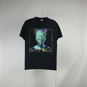 May include: Black t-shirt featuring a futuristic portrait. The graphic depicts a metallic figure with neon green and blue accents, wearing futuristic sunglasses. The design is set against a dark background.