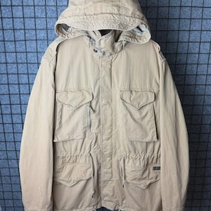 May include: A tan hooded jacket with multiple pockets and a drawstring waist. The jacket has a button and zipper closure and a high collar. The hood is adjustable and the jacket appears to be made of a durable, possibly water-resistant material.