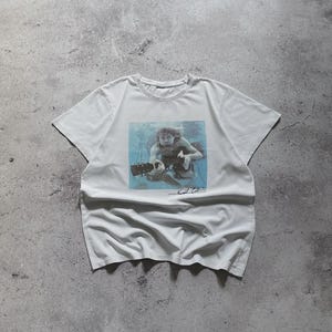 May include: White t-shirt featuring a square graphic of a person playing a guitar underwater. The graphic is in shades of blue and gray. The shirt has a crew neck and short sleeves. The artist's signature is below the graphic.