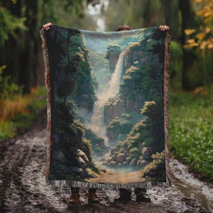 May include: A woven tapestry blanket featuring a scenic waterfall landscape with lush greenery and rocky terrain. The blanket has a fringed edge and is held up against a backdrop of a forest and a muddy path.