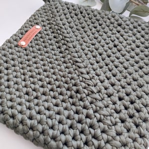 May include: A square, sage green crochet potholder with a loop for hanging. A small, rectangular leather tag with the words "Handmade, limited edition" is attached. The potholder is made of thick, textured yarn.