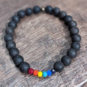 Puede incluir: Un brazalete de cuentas negras con un arcoíris de cuentas de colores. El brazalete está hecho de cuentas redondas negras con una sola fila de cuentas de colores en el centro.