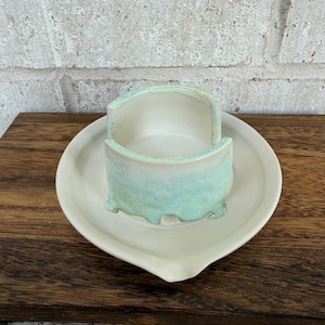 May include: A ceramic sponge holder featuring a circular base and a curved, open-front cylindrical top. The piece is finished in a smooth off-white glaze with a decorative, textured light green drip pattern around the exterior rim.
