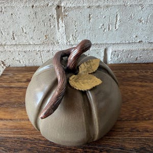 Pode incluir: Uma escultura de abóbora em cerâmica com um acabamento liso em verde-azeitona suave. O topo apresenta um caule texturizado castanho-escuro curvo e duas pequenas folhas douradas metálicas com um detalhe de superfície martelado.