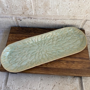 May include: An elongated oval ceramic tray featuring a light sage green glaze with a textured, repeating petal-like pattern radiating from the center. The surface has a glossy finish with subtle variations in color depth across the raised design.