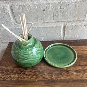 May include: A green ceramic essential oil diffuser with a round, textured body and a matching lid. The diffuser contains several wooden reeds. The items are displayed on a wooden surface against a white brick wall.