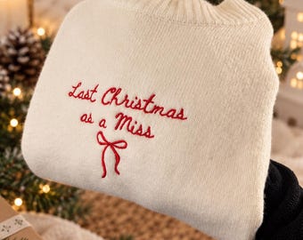 May include: A cream-coloured knit jumper featuring red embroidered text that reads "Last Christmas as a Miss" with a small red bow graphic underneath. The jumper has a ribbed crew neckline and a soft, textured knit fabric construction.