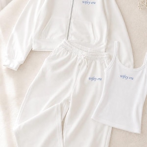 May include: A white zip-up hoodie, matching sweatpants with a drawstring waist, and a sleeveless tank top. Each piece features the text "wifey era" printed in a small blue font on the front. The set is made of a solid white fabric throughout.