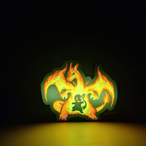 Può includere: Una luce illuminata a tema Charizard, con tonalità arancioni e gialle. Al centro, è visibile una silhouette di Charmander. È posta su una superficie di legno con uno sfondo scuro.