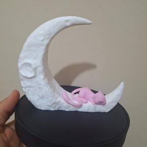 Puede incluir: Una escultura de luna creciente blanca con una superficie texturizada, apoyada sobre una base circular negra. Una pequeña figura de gato rosa está acurrucada durmiendo en la luna. Es un objeto decorativo.