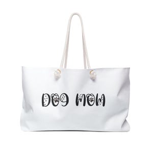 Puede incluir: Bolso tote blanco con asas de cuerda. Las palabras "DOG MOM" están impresas en negro, con huellas de patas que reemplazan la letra "O". Un accesorio elegante y práctico para los amantes de los perros.