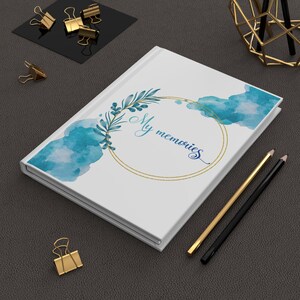 Puede incluir: Diario de tapa dura blanca con el texto "My memories" en escritura azul, rodeado por un círculo dorado y detalles en acuarela azul. También se ven lápices dorados y negros y clips dorados.