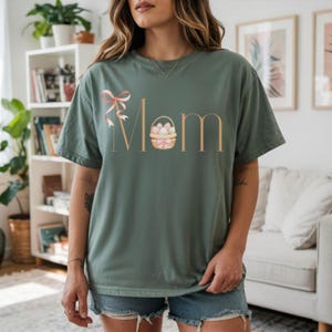 Puede incluir: Camiseta verde salvia con la palabra "Mom" en una fuente marrón claro. Un lazo rosa y una cesta de huevos de Pascua aparecen en el diseño. La camiseta es de corte holgado.